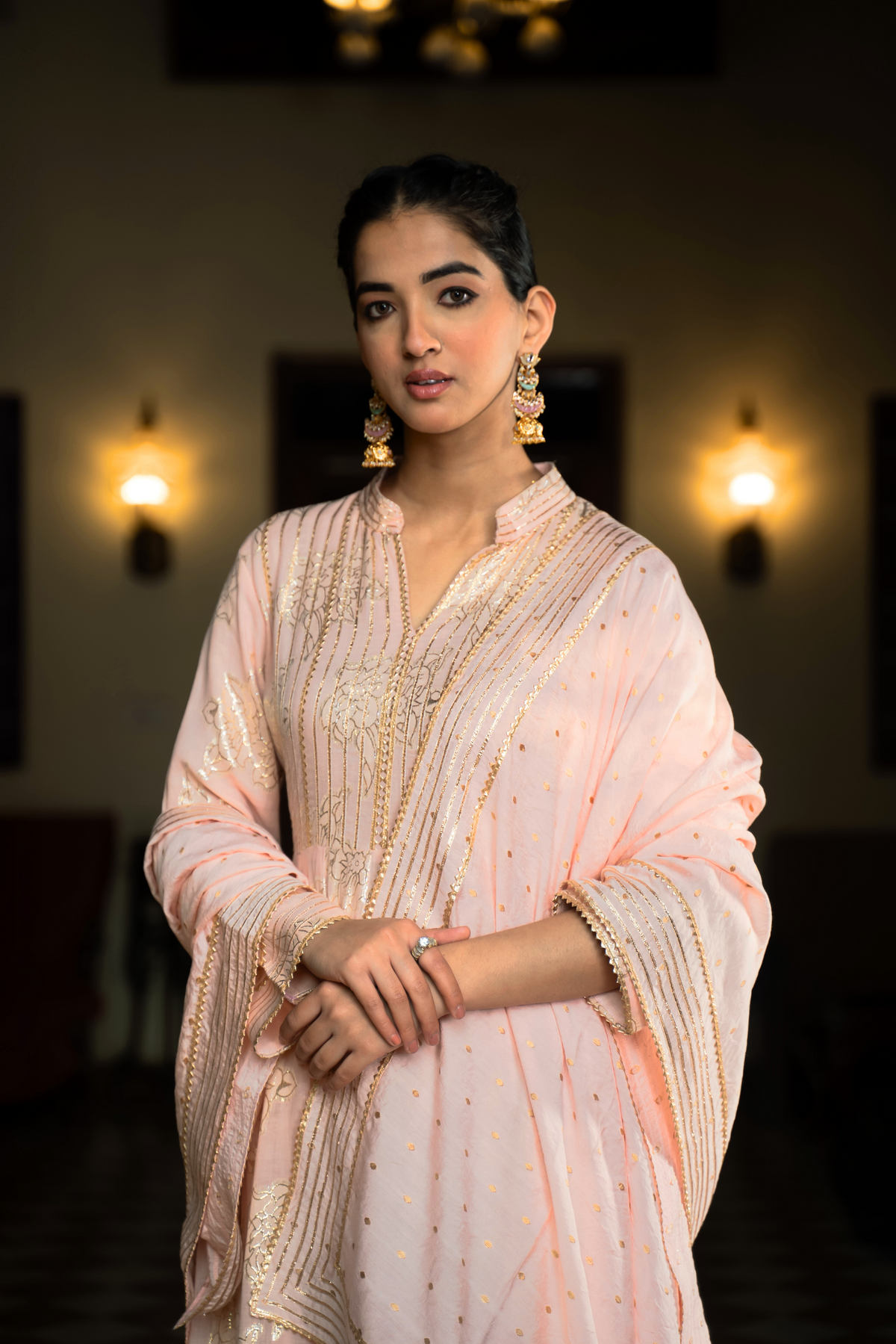 Closeup view of a  person stands elegantly in a pink embroidered anarkali kurta  set, surrounded by a traditional indoor setting with decorative lights and furnishings.