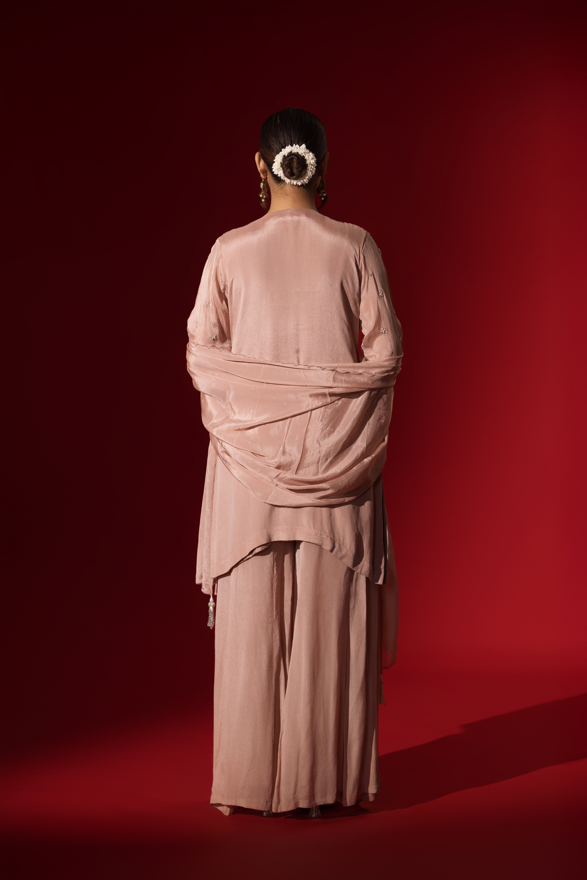 Back view of a woman wears an elegant pastel pink embroidered kurta set with intricate beadwork, standing against a vibrant red background.