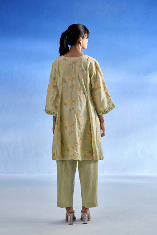 Back side of a woman is shown from the back wearing a green patterned tunic with three-quarter sleeves and matching olive green trousers, set against a blue gradient studio background.
