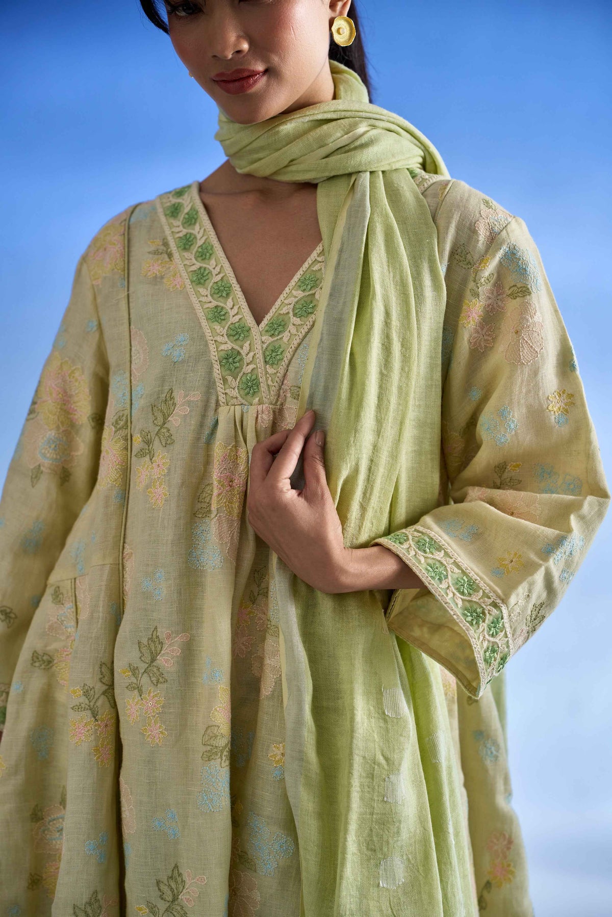 A woman wearing a pistachio green traditional outfit with floral embroidery, featuring a matching dupatta, set against a clear blue sky.