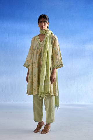 A woman modeling a light green traditional linen kurta set with a matching scarf, featuring a loose printed kurta, matching pants, and sandals, set against a blue gradient studio background.