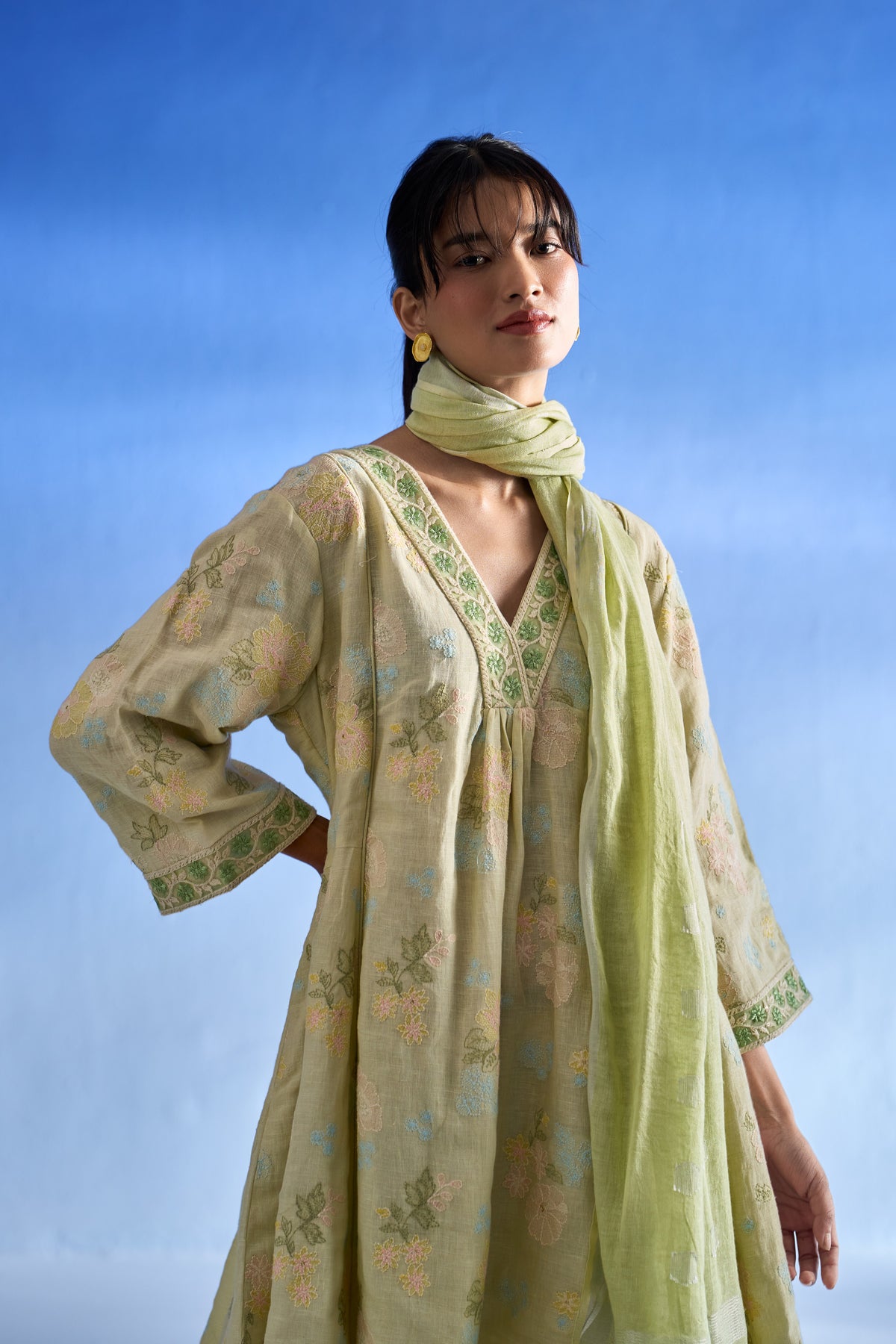 A woman modeling a pistachio green traditional kurta set with a matching scarf, featuring a soft floral pattern and green embroidered trim, set against a blue gradient background.