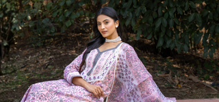 A woman wearing a pink and white printed traditional handblock printed kurta set with a white embroidered dupatta, seated outdoors in front of a leafy green background.