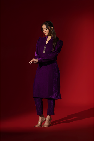 Side view of a woman in a vibrant purple velvet co-ord set with a feminine silhouette and elegant earrings, set against a rich red background.
