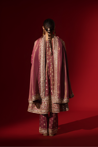 Back view of a woman stands elegantly in a rich maroon embroidered brocade kurta set adorned with intricate gold embroidery, against a deep red background.