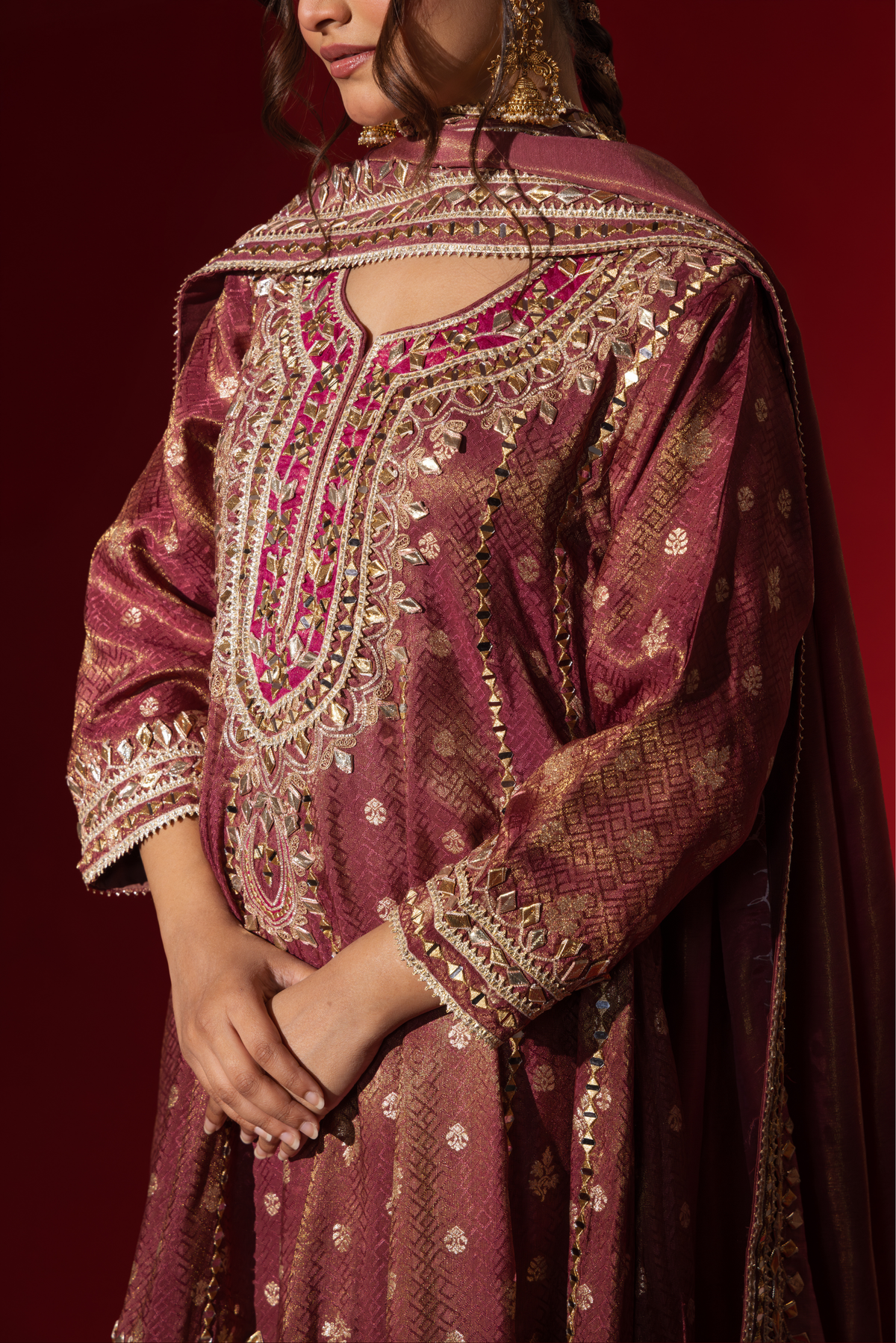 Closeup view of a woman's hand in a rich maroon embroidered brocade kurta set adorned with intricate gold embroidery, against a deep red background.