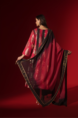 Back view of a woman poses in a vibrant red and black traditional outfit, adorned with intricate patterns and complemented by elegant jewelry.