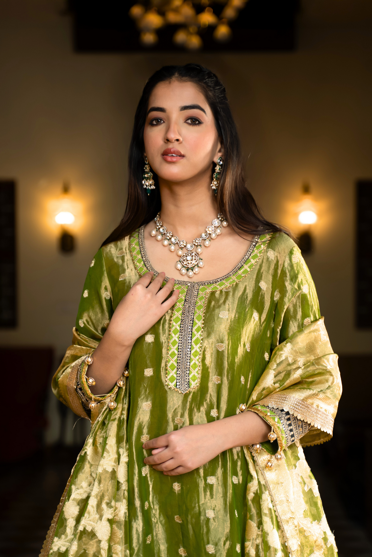 Closeup view of a woman in a traditional green anarkali outfit stands elegantly in a vintage-style room, surrounded by  decor and plush seating.