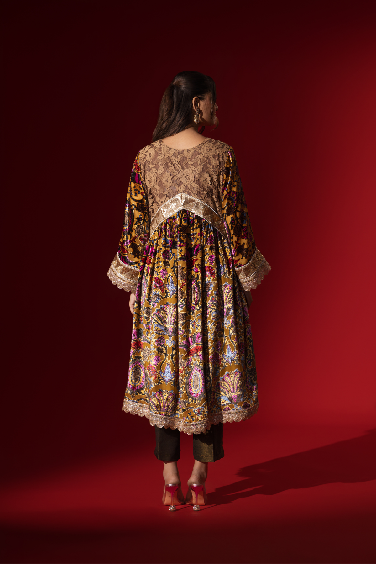 Back view of a woman stands confidently in a colorful, embellished regal printed velvet kurta set with a flowing golden dupatta against a vibrant red backdrop.