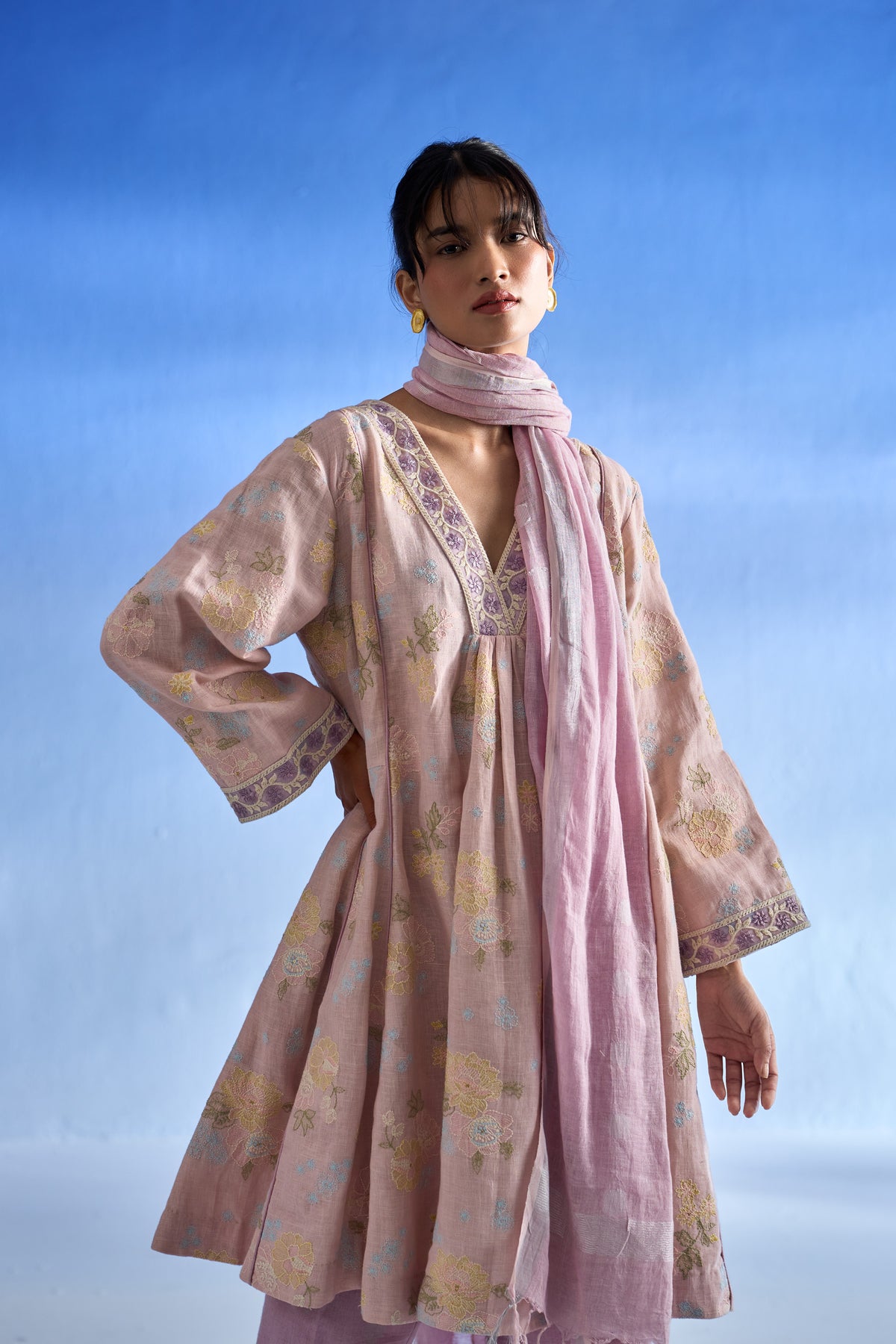 A woman modeling a light pink linen kurta set with a matching dupatta, featuring a loose tunic with floral patterns and embroidered details, set against a blue gradient background.