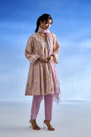 A woman models a traditional three-piece linen kurta set consisting of a light pink and beige floral printed kurta, matching trousers, and a coordinating pink dupatta, set against a blue gradient studio background.