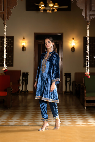Side view of a woman stands in a traditional setting, wearing a vibrant blue velvet co-ord set outfit with decorative accents, surrounded by elegant furnishings.