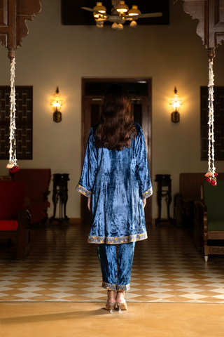 Back view of a woman stands in a traditional setting, wearing a vibrant blue velvet co-ord set outfit with decorative accents, surrounded by elegant furnishings.