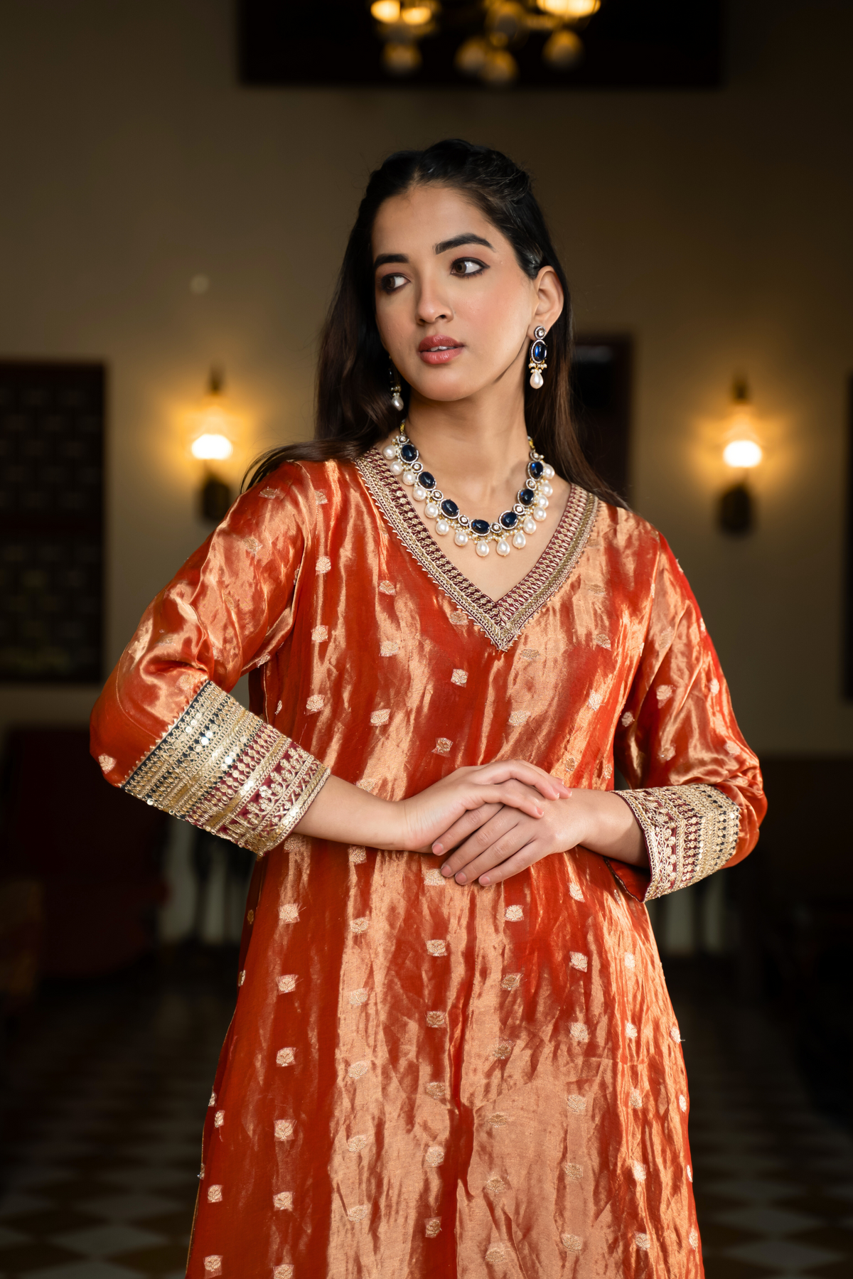 Closeup view of a  person in a traditional orange and golden banarsi silk kurta set outfit stands elegantly in a beautifully decorated room with vintage furnishings.