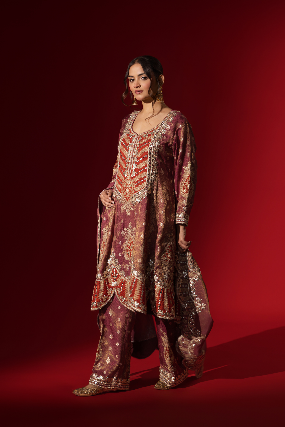 Side view of a woman wearing a richly embellished maroon embroidered brocade kurta set outfit with intricate gold detailing against a striking red background.