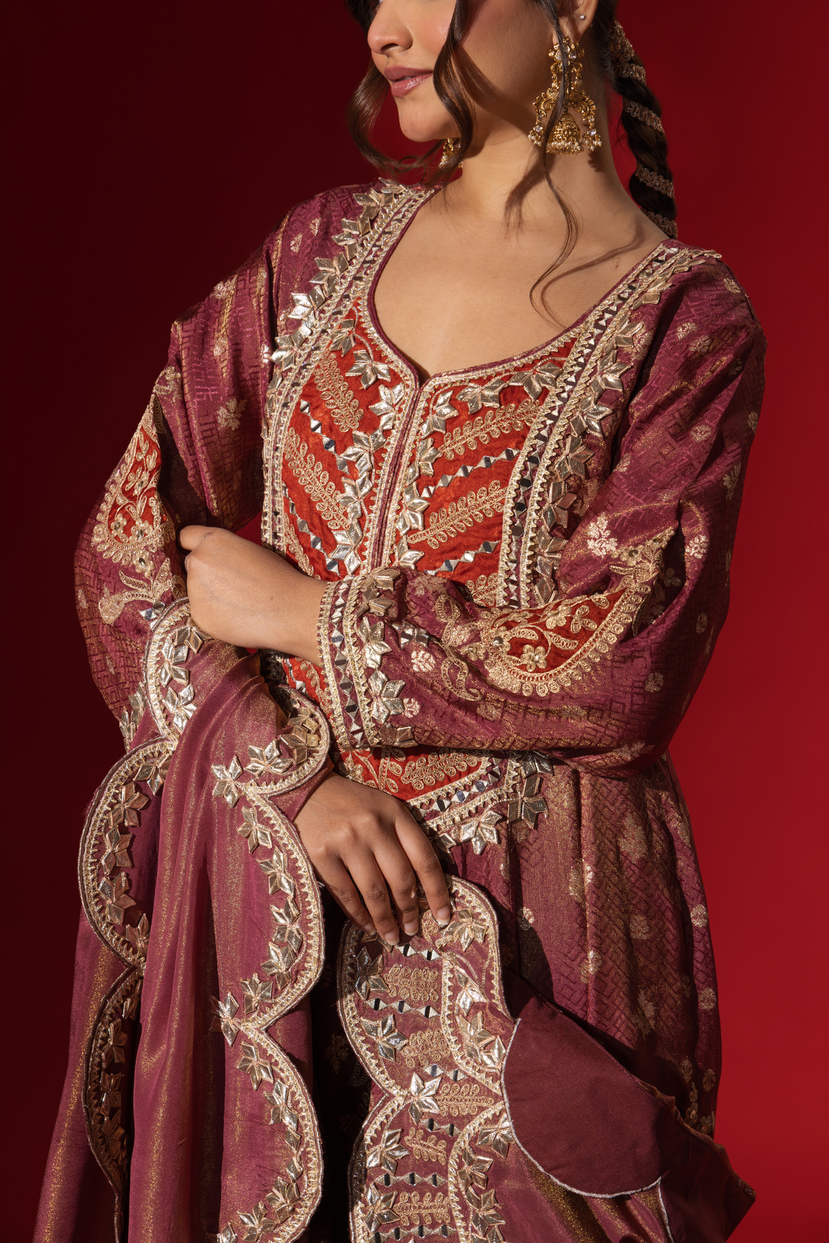 Closeup view of a woman's hand wearing a richly embellished maroon embroidered brocade kurta set outfit with intricate gold detailing against a striking red background.