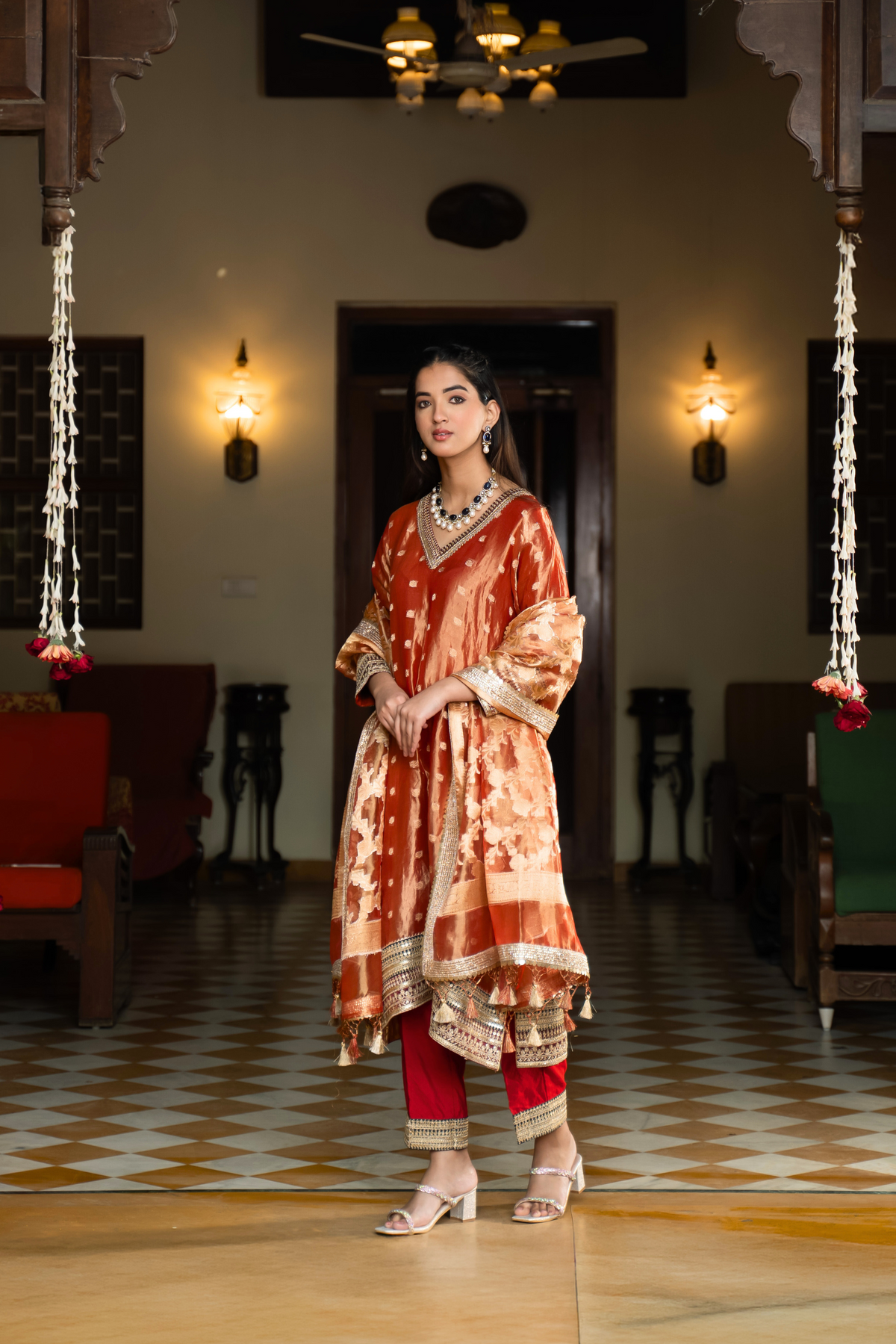 Side view of a  person in a traditional orange and golden banarsi silk kurta set outfit stands elegantly in a beautifully decorated room with vintage furnishings.