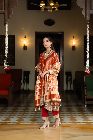 Side view of a  person in a traditional orange and golden banarsi silk kurta set outfit stands elegantly in a beautifully decorated room with vintage furnishings.