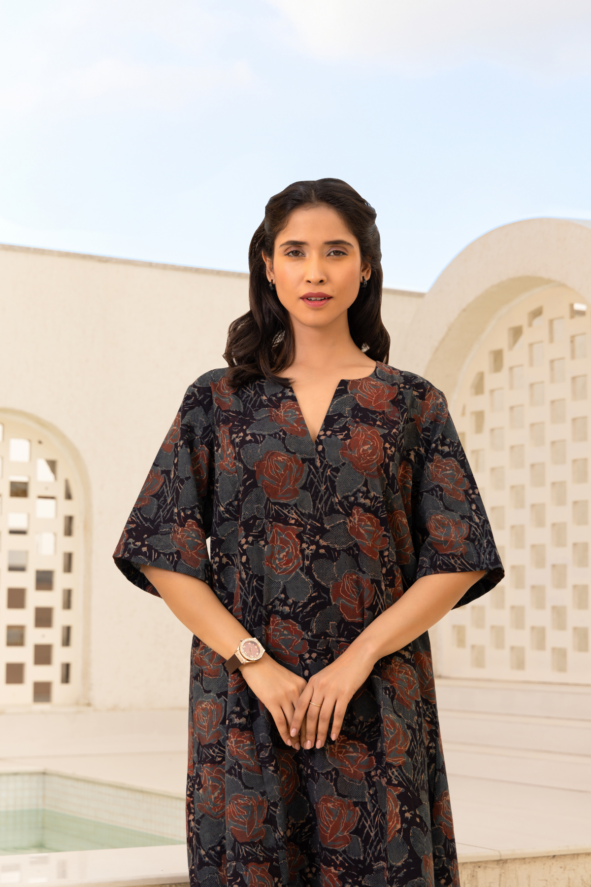 Closeup View of a woman wearing a dark floral-printed traditional outfit with a white hemline, paired with matching pants and black sandals, standing outdoors against a backdrop of light-colored architectural details and a blue sky.