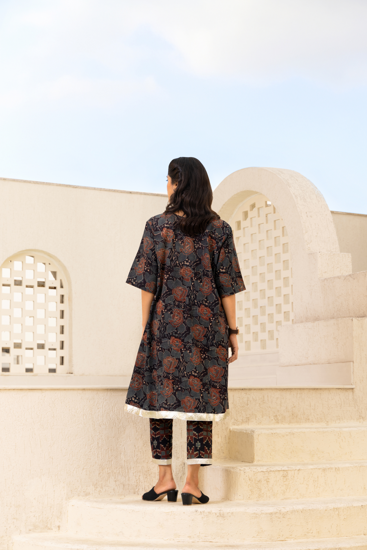 Back View of a woman wearing a dark floral-printed traditional outfit with a white hemline, paired with matching pants and black sandals, standing outdoors against a backdrop of light-colored architectural details and a blue sky.