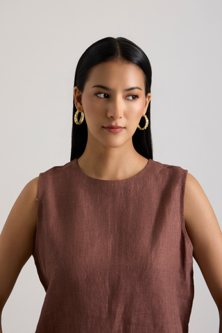 A woman with long dark hair wearing a sleeveless dusty rose-brown top and gold hoop earrings, photographed against a plain light gray studio background.