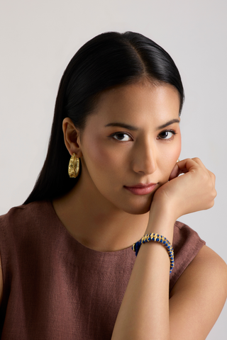 A woman with long black hair posing in a brown linen top, wearing a gold hoop earring and a blue and gold beaded bracelet against a light gray background.