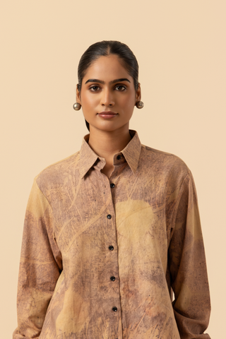 Closeup view of a model stands against a light beige background, wearing a patterned co-ord set which has button-up shirt and wide-legged beige pants, accessorized with heels.