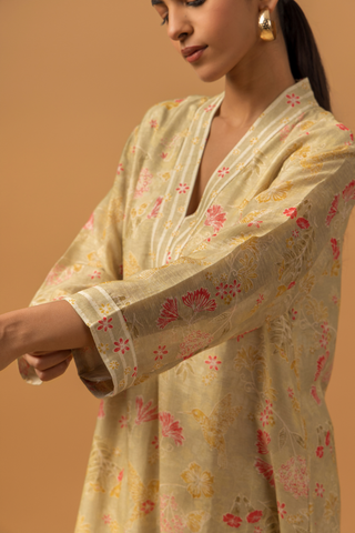 A woman in a floral-patterned, long beige linen co-ord set and striped pants, showcasing hand sleeves, stands against a warm beige background.