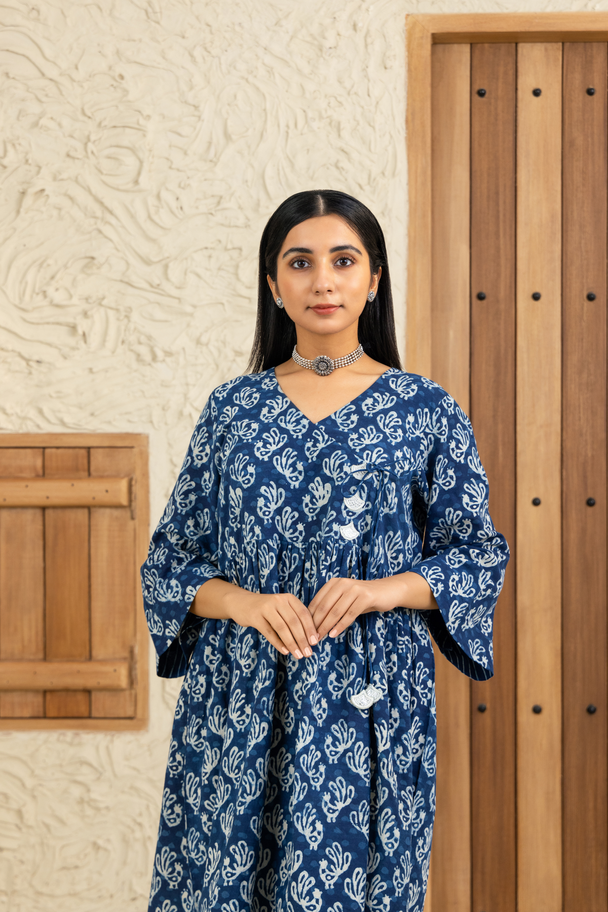 Closeup view of a woman wearing a blue and white printed co-ord set with a V-neck and tassel details, standing against a wooden and textured beige background.