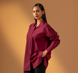 A woman modeling a long-sleeve button-up shirt in a deep burgundy or maroon color, paired with black pants, set against a warm, solid gradient studio background.