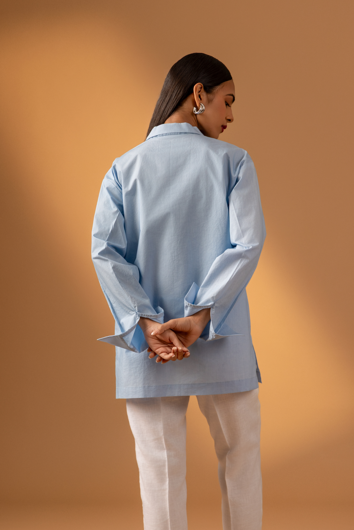 Back view of a women stands confidently wearing a light blue oversized shirt, white pants, and stylish sandals, against a warm beige background.