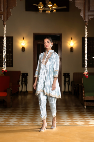 Side view of a woman wears a light blue ethnic velvet co-ord set outfit, standing gracefully in a spacious, elegantly decorated room with warm lighting and traditional furniture.
