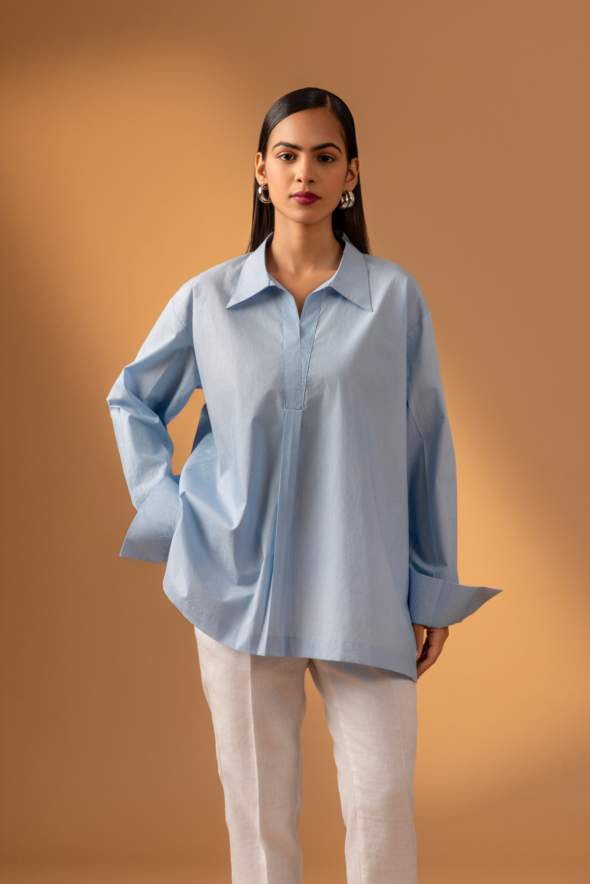 A women stands confidently wearing a light blue oversized shirt, white pants, and stylish sandals, against a warm beige background.