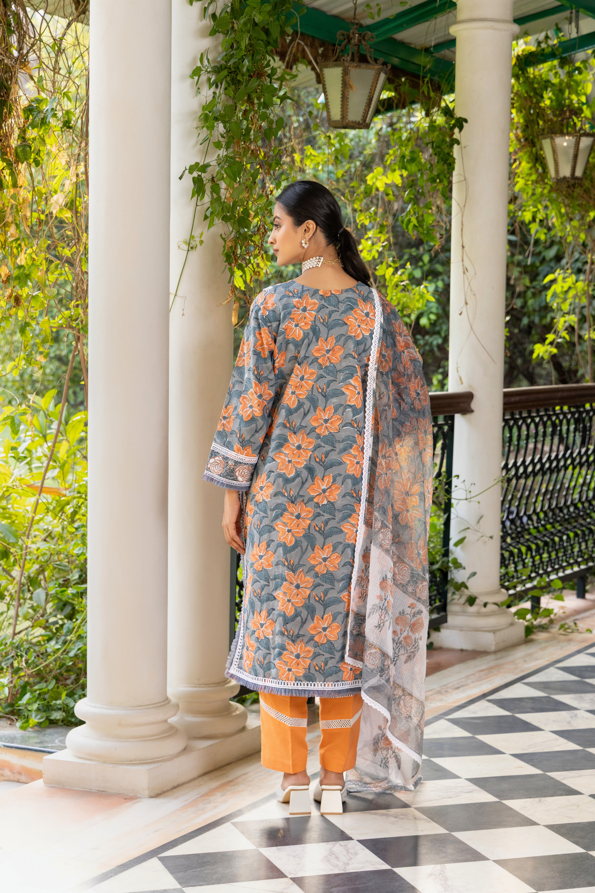 Back view of a woman stands elegantly in a floral, gray and orange cotton kurta set, complemented by a sheer scarf, amidst a lush green background.
