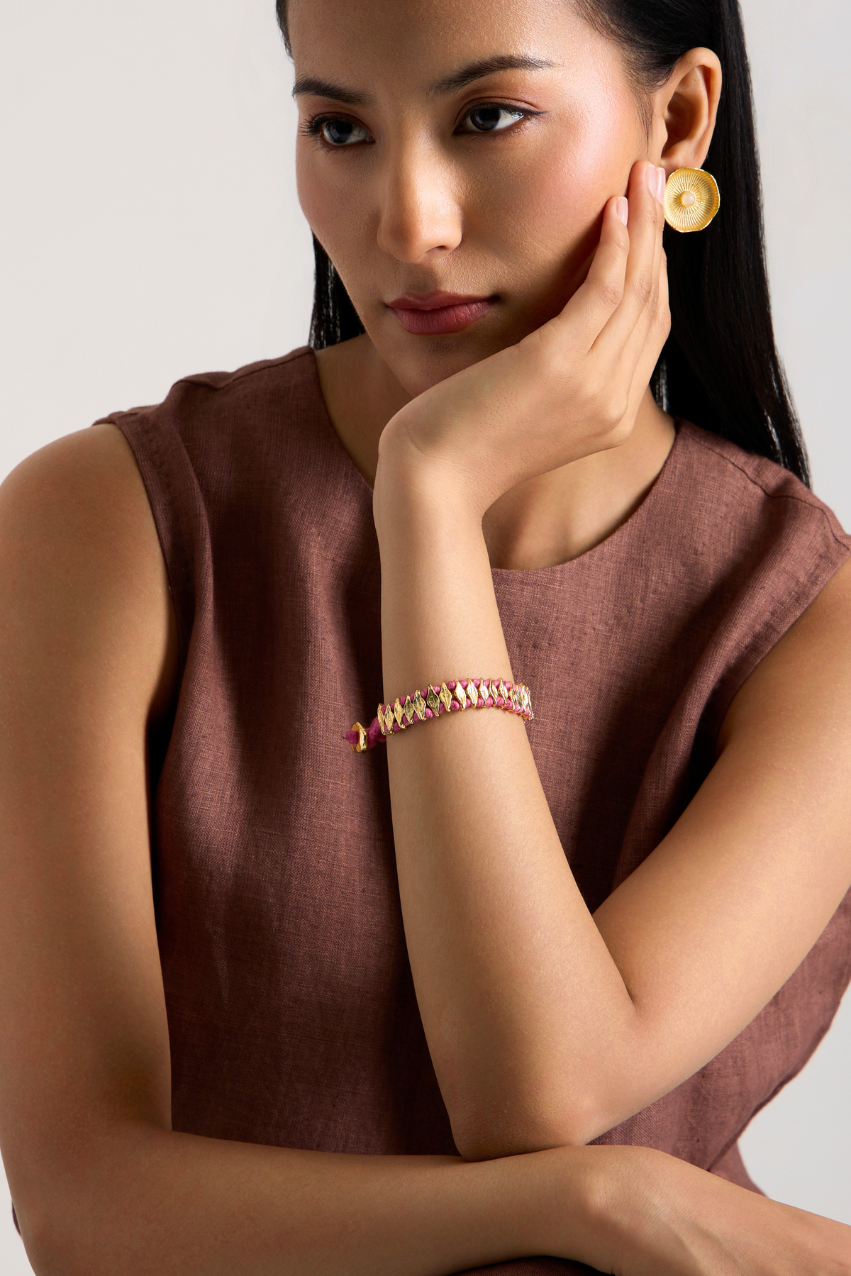 A woman wearing a delicate gold chain bracelet with pink and white beads and a matching gold disc earring, posing in a sleeveless brown co-ord set against a light gray background.