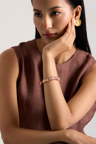 A woman wearing a delicate gold chain bracelet with pink and white beads and a matching gold disc earring, posing in a sleeveless brown co-ord set against a light gray background.