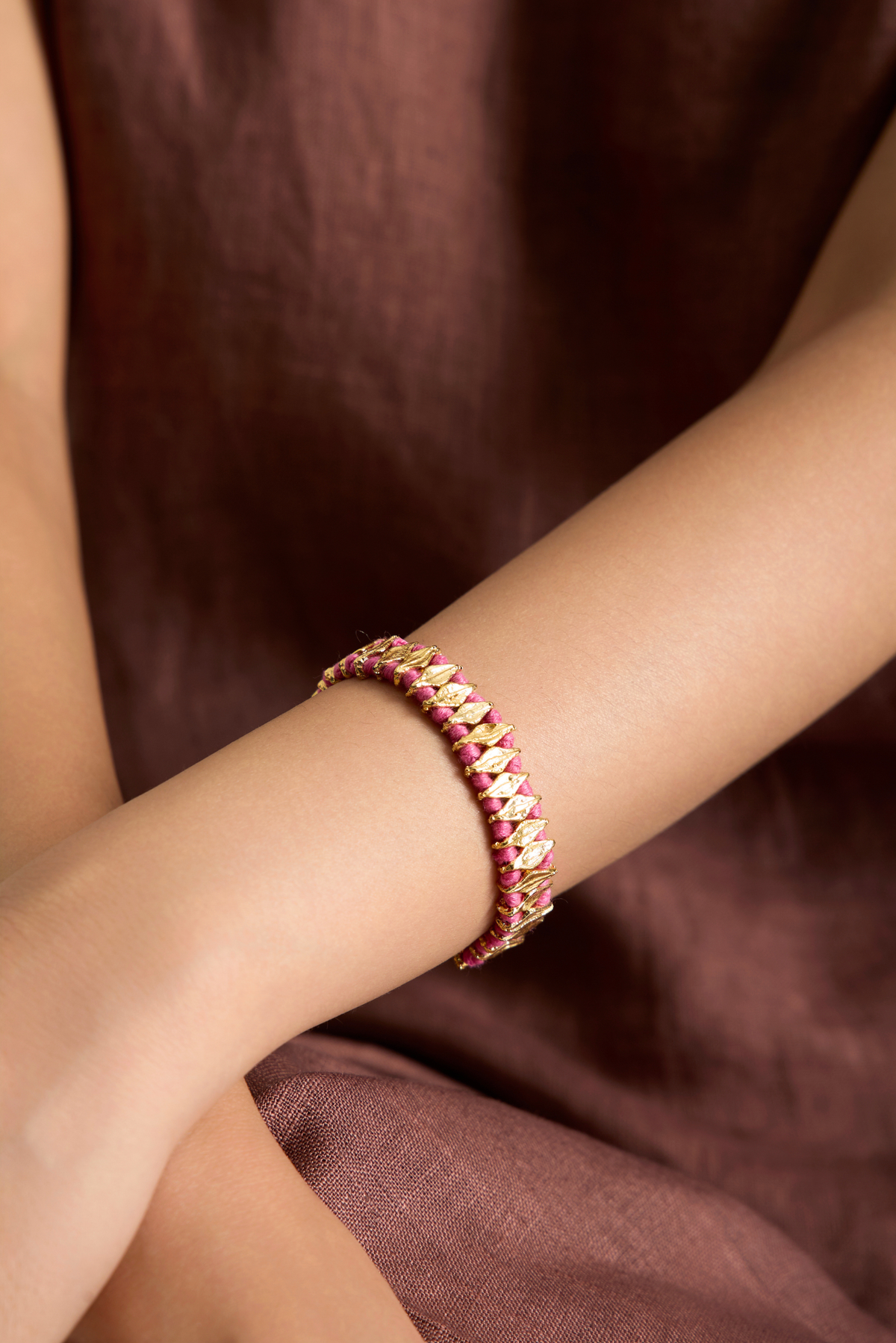 A close-up of a person's wrist wearing a delicate gold chain bracelet adorned with small pink gemstones, set against a soft brown fabric background.