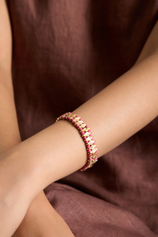 A close-up of a person's wrist wearing a delicate gold chain bracelet adorned with small pink gemstones, set against a soft brown fabric background.
