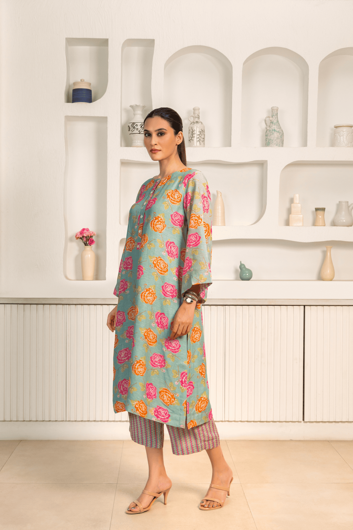 A woman is dressed in a light blue kurta with vibrant pink and orange floral prints, paired with patterned pants and posing against a white minimalist backdrop featuring built-in shelves with vases and decorative items.