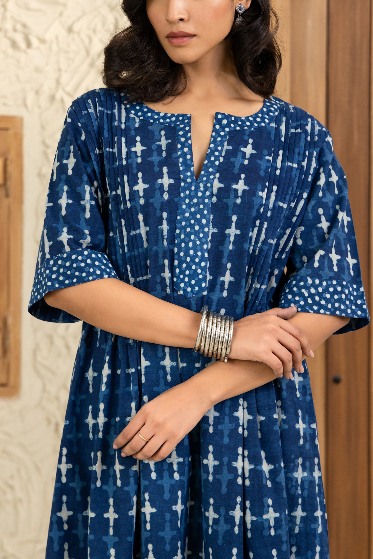 Closeup view of a model with shoulder-length wavy hair wears a blue printed co-ord set with white patterns, silver bangles, and earrings. She stands in front of a wooden door and textured beige wall, gazing softly at the camera.
