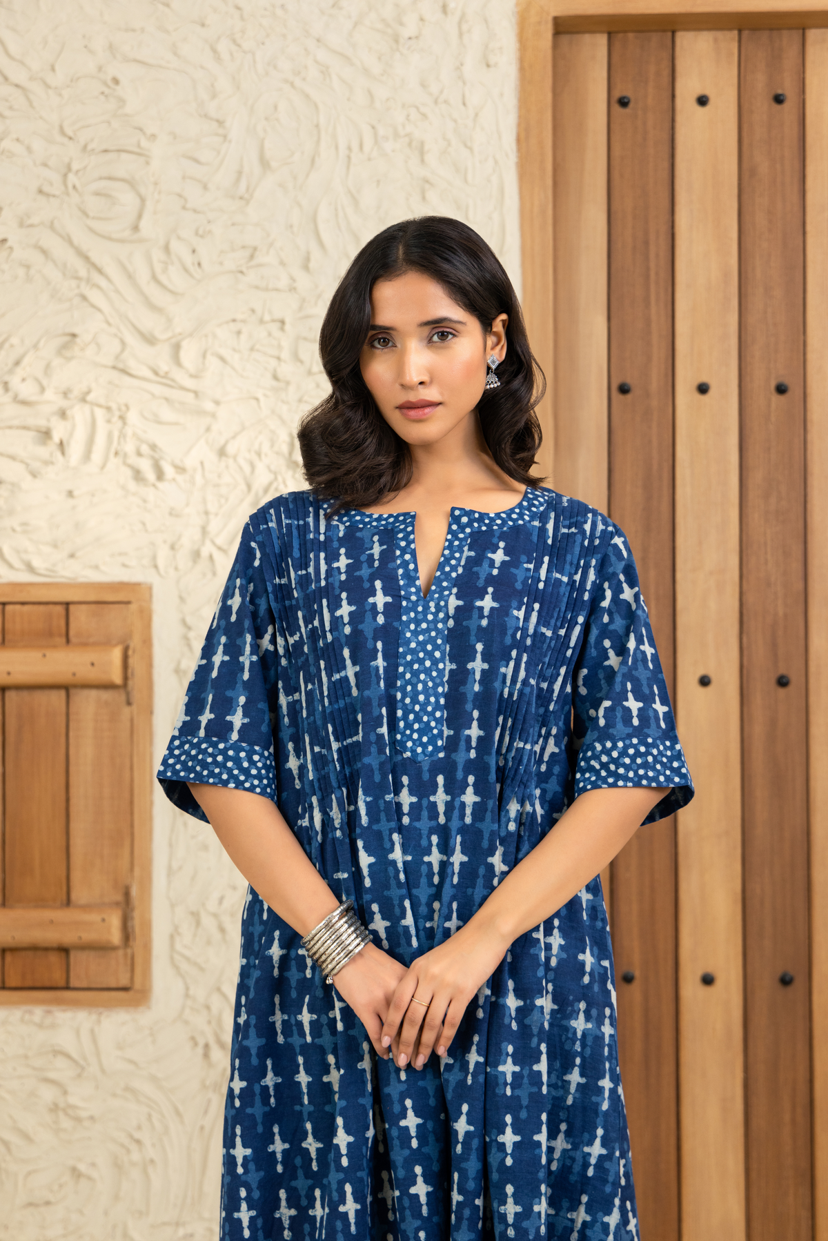 Closeup view of woman with shoulder-length wavy hair wears a blue printed co-ord set with white patterns, silver bangles, and earrings. She stands in front of a wooden door and textured beige wall, gazing softly at the camera.