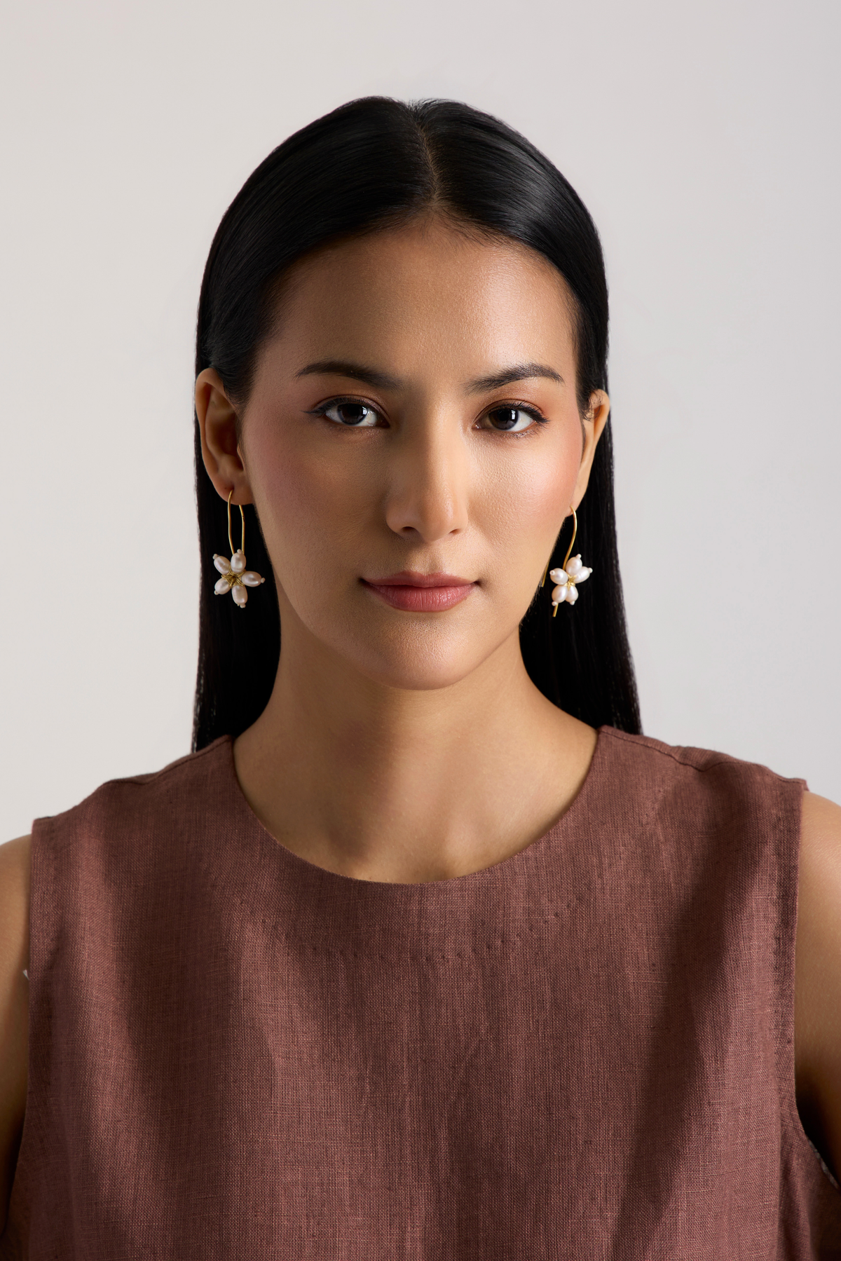 A woman with long black hair wearing a sleeveless brown top and gold flower-shaped dangle earrings, photographed against a plain light gray background.