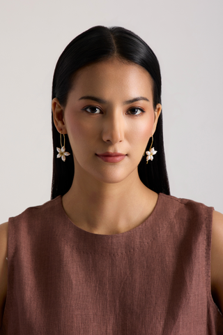 A woman with long black hair wearing a sleeveless brown top and gold flower-shaped dangle earrings, photographed against a plain light gray background.