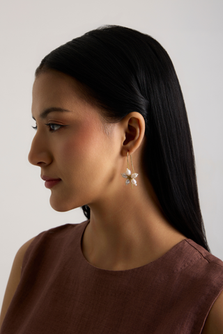 A woman in profile wearing a dangling flower-shaped earring with white petals and a gold center, paired with a brown sleeveless top against a plain light gray background.