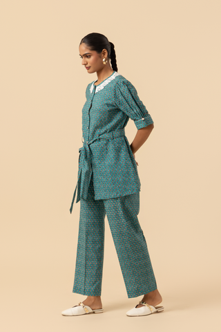 Side view of a person stands confidently, wearing a teal patterned outfit with a cinched waist and a collar, set against a neutral background.