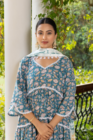 Closeup view of a woman stands gracefully in a patterned blue kurta set and white pants, in a lush green setting with ornate pillars and checkered flooring.