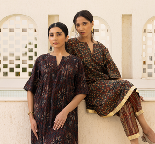 Two women modeling traditional South Asian ajrakh co-ord set in front of a cream-colored stucco wall with arched windows. One wears a dark brown tunic with a blue and beige floral pattern, and the other wears a matching brown kameez with striped pants featuring gold fringe.