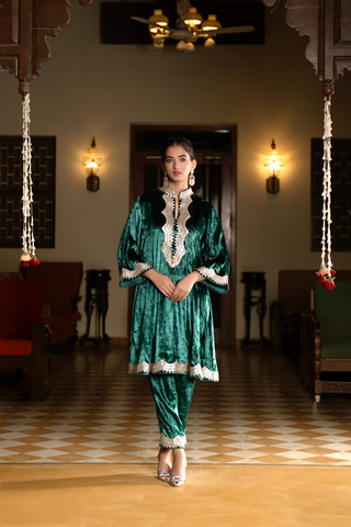 A woman in a green velvet co-ord set traditional outfit stands in a warmly lit room with decorative lighting and patterned flooring.