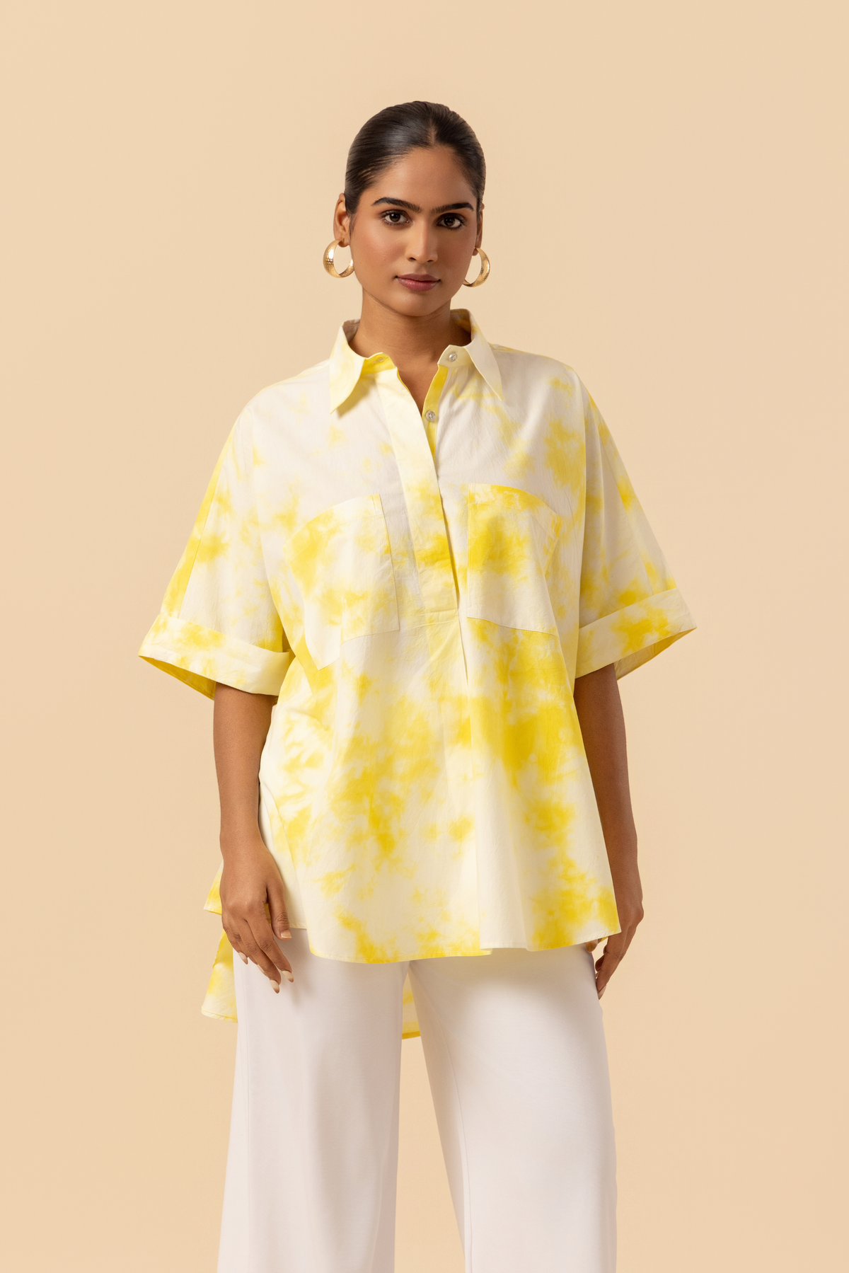 Closeup view of a model poses confidently in a yellow tie-dye oversized shirt and white wide-leg pants against a soft beige background.
