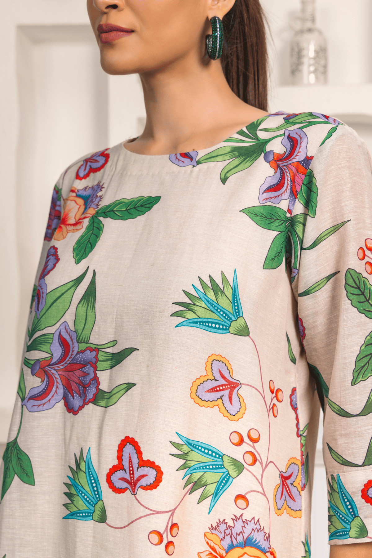 Closeup view of  woman is wearing a light-colored floral-printed kurta set paired with patterned pants. She is posing against a minimalist white background featuring built-in shelves decorated with vases and small decorative items.